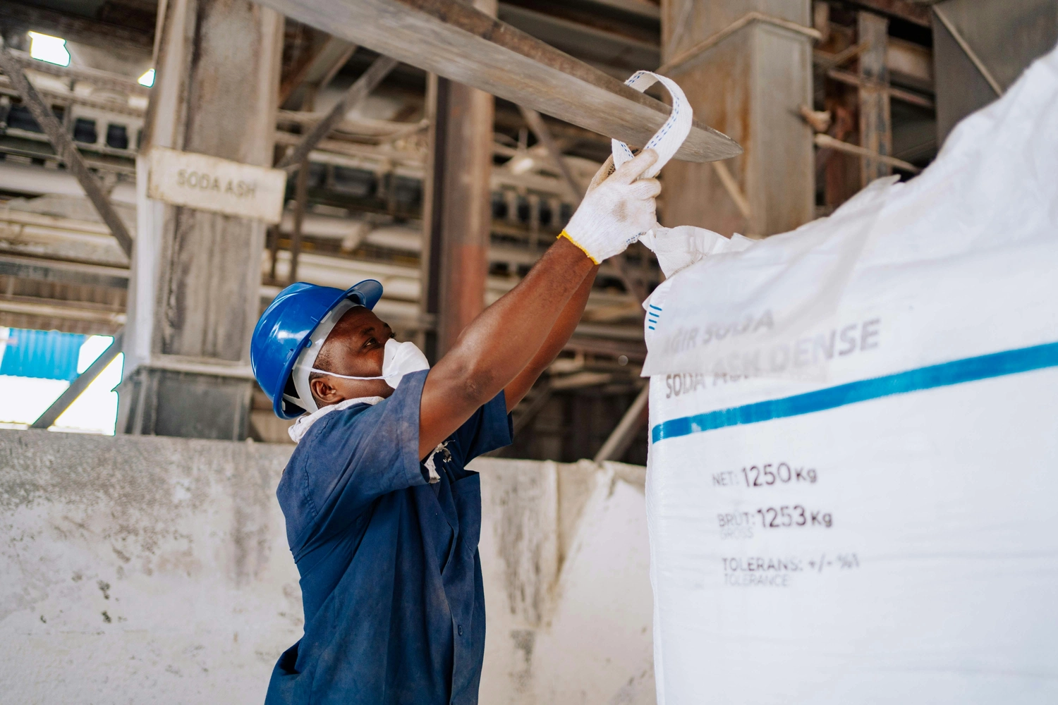 Solid Cargo GmbH - Internationalen Handel - Ein Arbeiter mit blauem Schutzhelm und Atemschutzmaske sichert einen großen weißen Sack mit Transportgütern.